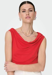 Haut sans manches rouge avec un col drapé. Fabriqué à partir d'un tissu lisse, il présente une coupe décontractée et un léger éclat. Le modèle pose devant un fond neutre.