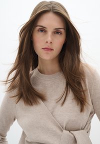 Beige Strickpullover mit Wickeldesign und seitlichem Bindeband. Besitzt einen Rundhalsausschnitt und eine weiche Textur. Das Modell hat lange, glatte Haare.