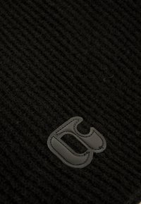 Bonnet en tricot noir avec une finition texturée et un patch en cuir noir proéminent, arborant une lettre stylisée.
