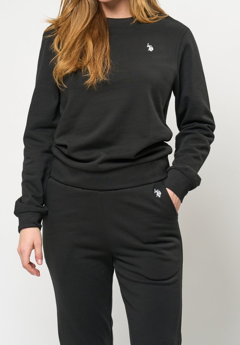 Femme portant un sweat-shirt et un pantalon noirs avec un petit logo blanc brodé sur la poitrine et la poche, main dans la poche, fond neutre.