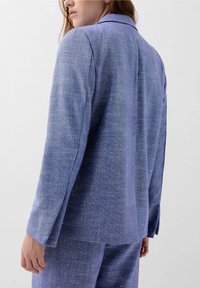 Blauwe textuur blazermantel met een getailleerde pasvorm, voorzien van een reverskraag en subtiele metallic glitters verspreid over de stof. Lange mouwen, rechte snit.