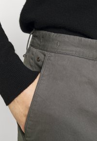 Ręka w przedniej kieszeni szarych spodni, ubrana w czarny sweter z długim rękawem, pokazująca teksturę materiału i szczegóły pasa.