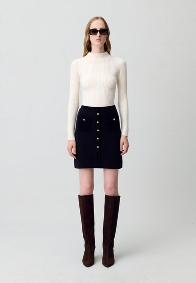 Maglione a collo alto a costine color crema, abbinato a una gonna blu scuro con tasche frontali e bottoni dorati, completato da stivali marroni sopra il ginocchio.