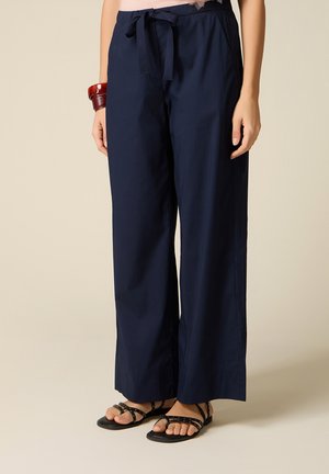 Marineblå bukser med vide ben og snøring i taljen, båret med sorte sandaler med nitter og røde armbånd på en hævet arm mod en beige baggrund.