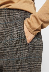 Pantalons à motifs marron et noir avec des accents bleus, dotés d'une poche latérale et d'une finition texturée. Un pull beige est partiellement visible.