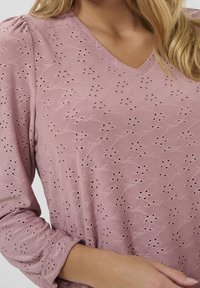 Maglietta a maniche lunghe con scollo a V in rosa polveroso, con ricami floreali in rilievo e dettagli in pizzo. Tessuto morbido con una leggera lucentezza e maniche arricciate.