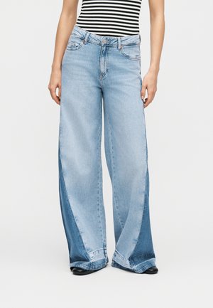 Femme portant un jean taille haute à jambes larges avec des panneaux bleu clair et bleu foncé, ainsi que des chaussures noires à bouts pointus, assortis à un haut rayé noir et blanc.