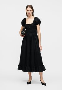 Γυναίκα που φοράει μαύρο μεσάτο φόρεμα με φουσκωτά μανίκια και βολάν στο τελείωμα, κρατώντας μαύρη τσάντα και φοράει μαύρες μυτερές γόβες.