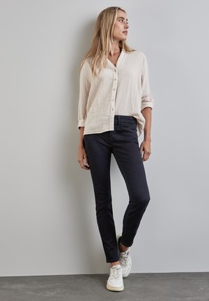 Camisa de botones en crema con tejido texturizado, combinada con pantalones ajustados de un azul marino oscuro. El modelo lleva zapatillas blancas. Diseño simple y limpio.