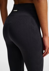 Czarne legginsy z wysokim stanem o prążkowanej fakturze, z delikatnym wzorem jodełki i małą metką z logo na tylnej części pasa.