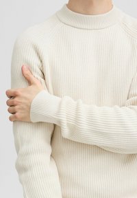 Maglione di colore crema lavorato a maglia con una texture a coste, polsini aderenti e scollatura rotonda. Il tessuto sembra morbido e leggero.