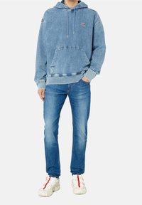 Sudadera de mezclilla en azul claro con un bolsillo frontal tipo canguro, puños acanalados, cordones ajustables y un pequeño parche de logo rojo. Combinada con jeans azules y zapatillas blancas.