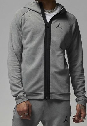 Mężczyzna w szarej bluzie Nike Jordan z kapturem i dopasowanych dresach z czarnym zamkiem oraz logo Jumpman, pokazuje pierścionki i tatuaże na dłoniach.