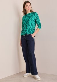 Grön mönstrad tröja med långa ärmar, ihop med mörka jeansbyxor. Bärs med vita sneakers, mot en neutral bakgrund.