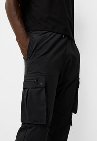 Svarta cargo-byxor i lätt tyg, med flera fickor, en ficka med dragkedja på sidan och ett elastiskt midjeband.