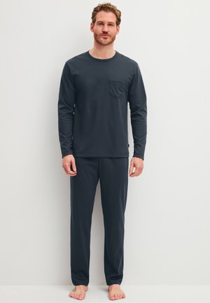 Man staat op blote voeten, draagt een donker langwerpig shirt met zakje en bijpassende broek tegen een effen lichte achtergrond.