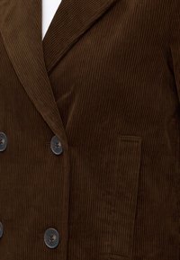 Veste en velours côtelé marron foncé avec larges revers, quatre boutons devant et une poche plaquée rectangulaire sur le côté droit.
