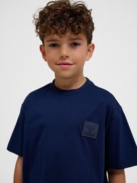 Junge mit lockigem braunem Haar, der ein marineblaues T-Shirt mit einem strukturierten Logo-Patch auf der Brust trägt, steht vor einem einfarbigen hellen Hintergrund.