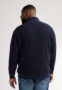 Marinblå texturerad tröja med hög krage, stickad mönster och ribbade ärmslut, kombinerad med blå jeans. Bakifrån visad.