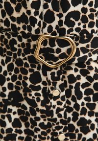 Close-up van luipaardprintstof met een gouden gesp in onregelmatige vorm en zichtbare knopen in het midden.