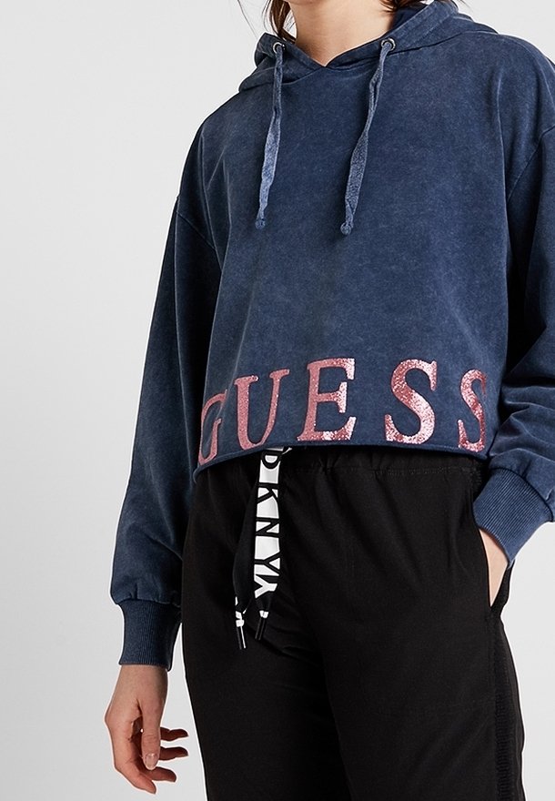Personne portant un sweat à capuche bleu court avec un lettrage rose pailleté « GUESS » et un pantalon noir avec des cordons blancs à motifs.