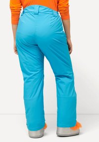 Blauwe skibroek met een gladde, glanzende textuur, hoge tailleband en wijde pijpen. Oranje bontaccenten op grijze schoenen.