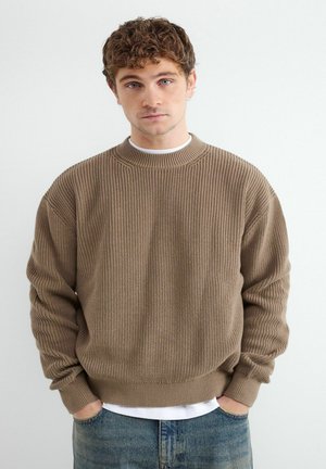 Młody mężczyzna o kręconych brązowych włosach, ubrany w brązowy sweter prążkowany i niebieskie dżinsy, stoi z rękami w kieszeniach na białym tle.