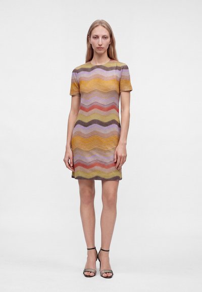Femme debout portant une robe courte à manches courtes, longueur genou, avec des rayures ondulées multicolores et des sandales à talons hauts noires à bride cheville.