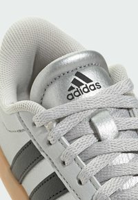 Sive atletske cipele s teksturiranim kožnim gornjim dijelom, srebrnim detaljima, tri crne pruge i svijetlosivim vezicama. Vidljiv logo proizvođača.