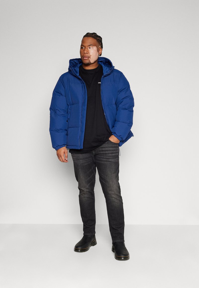 Giacca a vento blu con cappuccio, chiusura con zip e polsini elastici, indossata sopra una maglietta nera, abbinata a jeans scuri e stivali neri.