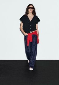 Schwarzes kurzärmeliges Oberteil mit Knöpfen, roter Pullover in der Taille gebunden und lockere dunkelblaue Jeans. Das Model trägt große Sonnenbrillen und weiße Schuhe.