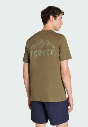 Mann trägt ein olivgrünes TERREX-T-Shirt mit Berglogo auf dem Rücken und marineblaue Shorts, seitlich stehend vor einfachem Hintergrund.
