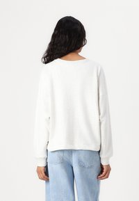Felpa bianca oversized con spalle scese e scollatura a girocollo. Presenta una texture morbida e una vestibilità comoda. Indossata con jeans azzurri.