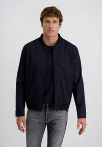Man met kort krullend haar draagt een marineblauwe jas over een donker shirt en grijze jeans, staand tegen een effen lichte achtergrond.