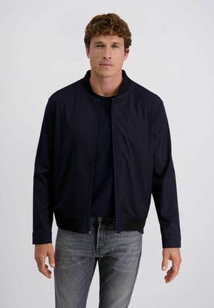 Mann mit kurzem lockigem Haar, der eine marineblaue Jacke über einem dunklen Hemd und grauen Jeans trägt und vor einem schlichten hellen Hintergrund steht.