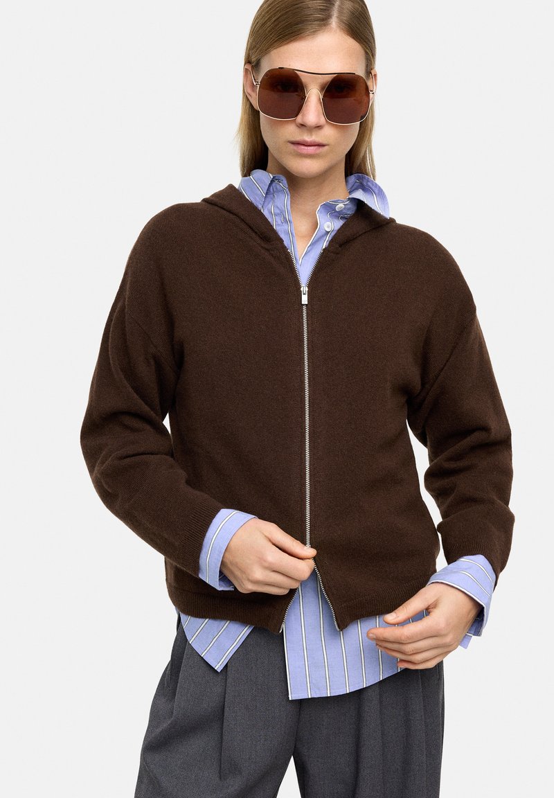 Brązowy polarowy hoodie zapinany na zamek, wykonany z miękkiego materiału, z kapturem i luźnym krojem, noszony na jasnoniebieskiej koszuli w paski. Model ma na sobie okulary przeciwsłoneczne.