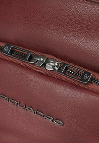 Bolso de cuero rojo que presenta una cremallera prominente con un tirador de metal y un logo plateado "Piquadro" grabado en la superficie. Textura suave.