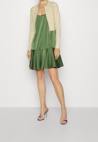 Giacca bomber in pizzo beige sopra un vestito satinato verde con orlo svasato, abbinato a tacchi alti a strappi viola.