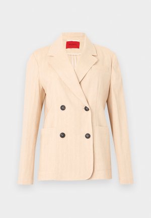 Blazer beige croisé avec revers crantés, quatre boutons noirs, deux poches plaquées à l'avant et une étiquette intérieure rouge.