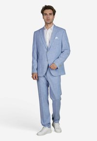 Man in een lichtblauw pak met een wit shirt en pochet, die zelfverzekerd staat met witte sneakers op een effen achtergrond.