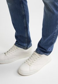 Weiße Schnür-Sneaker, getragen mit blauen Denim-Jeans von einer Person, die auf einem weißen Hintergrund steht.