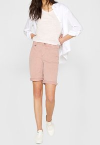 Pantaloni corti di cotone rosa chiaro con orli arrotolati, abbinati a una camicia bianca a bottoni e una canotta beige, completati da sneakers bianche.