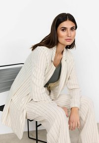 Beige gestreifter Anzug aus strukturiertem Stoff, mit einem taillierten Blazer und weiteren Hosen, kombiniert mit einem dunkelgrünen Oberteil.
