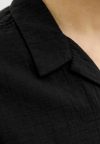 Camisa de lino negra con un cuello estructurado y una superficie texturizada. La tela parece ligera y tiene detalles de costura sutiles.