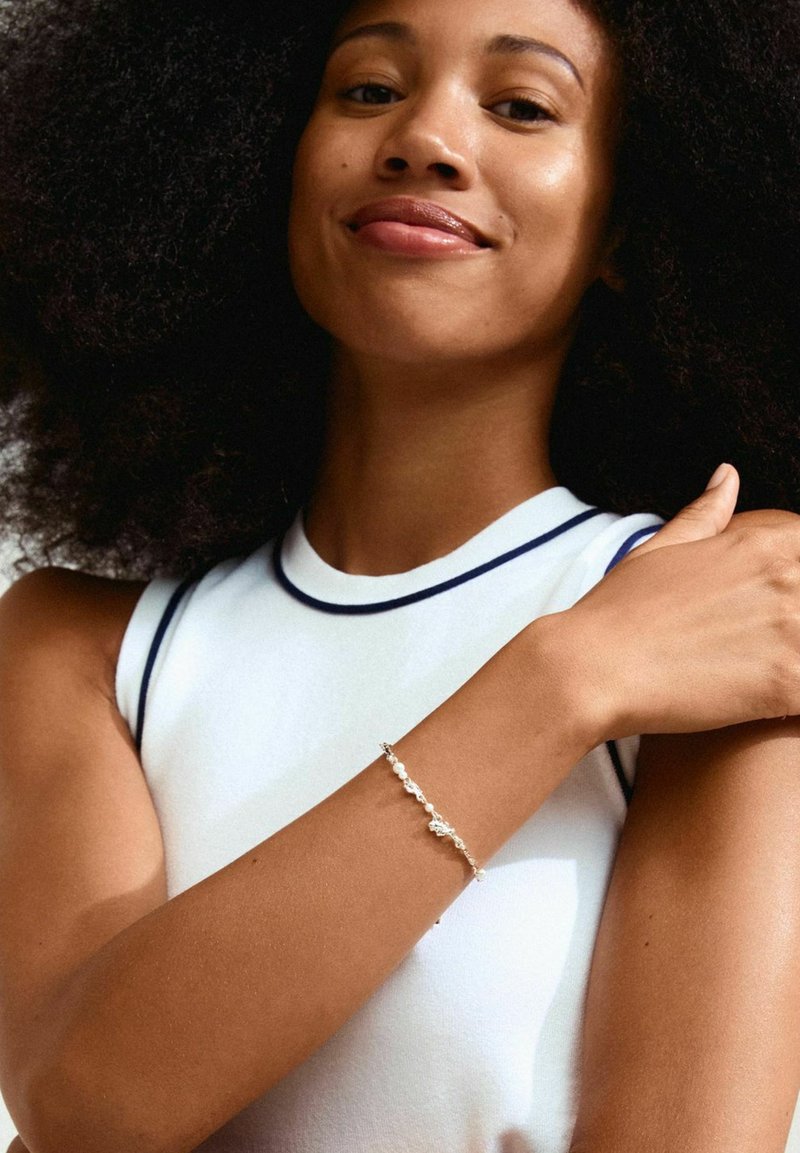 Silbernes Armband mit kleinen Perlen, das am Handgelenk eines Models getragen wird. Das Model trägt ein weißes ärmelloses Oberteil mit marineblauen Akzenten.