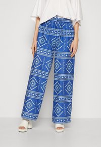 Un pantalon bleu à motifs avec des motifs géométriques blancs, coupe ample, taille élastique et lien. Porté avec des sandales à talons carrés blanches.
