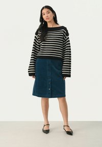 Gestreifter schwarz-weißer Strickpullover mit weitem Halsausschnitt und überschnittenen Schultern, kombiniert mit einem blauen Cord-Midi-Rock mit Knöpfen auf der Vorderseite.