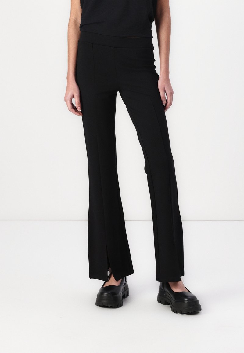 Femme portant un pantalon noir ajusté avec des fentes à l'avant de l'ourlet et des chaussures noires épaisses sur un fond blanc uni.