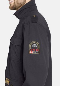 Black jacket featuring a tall collar, shoulder epaulets, and multiple pockets. Notable embroidered patch on the sleeve with mountain design.