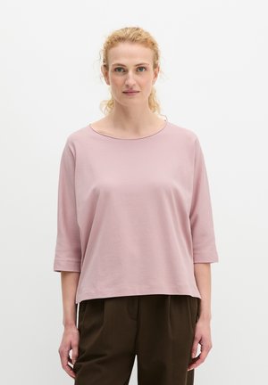 Kobieta o kręconych blond włosach, ubrana w luźną, jasnoróżową bluzkę z rękawami do łokcia oraz ciemnobrązowe spodnie, stojąca na jednolitym białym tle.